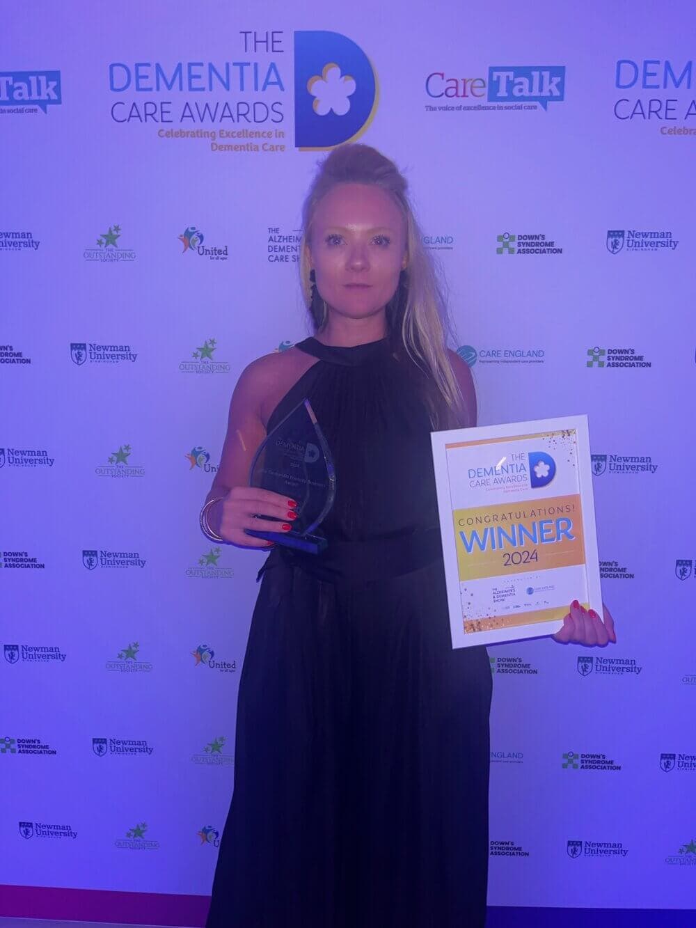 A woman holding a trophy and a winner's certificate at the Dementia Care Awards, standing in front of a sponsor backdrop. - Home Instead