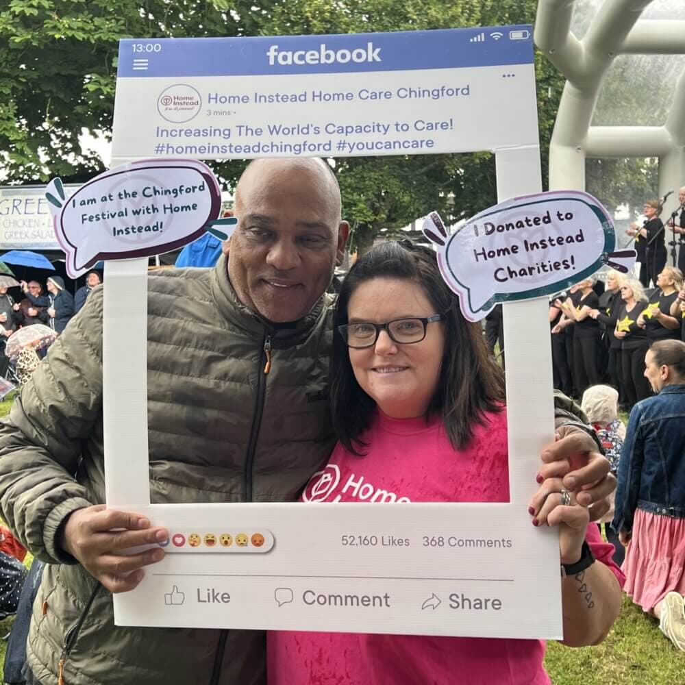 Two people posing with a Facebook frame sign promoting Home Instead Care at Chingford Festival, surrounded by outdoor crowd. - Home Instead