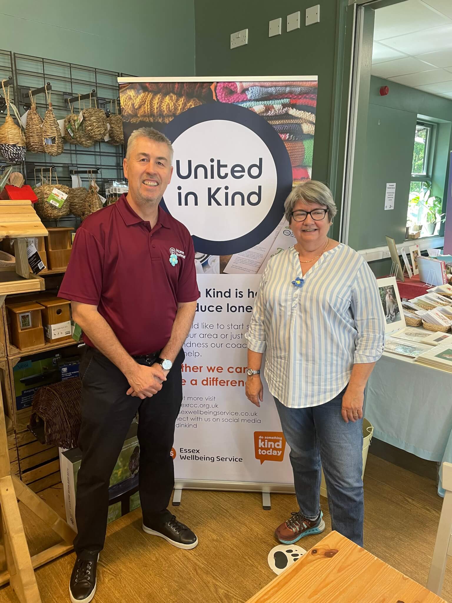 Two people stand smiling in front of a "United in Kind" sign in a room decorated with crafts and wellness products. - Home Instead