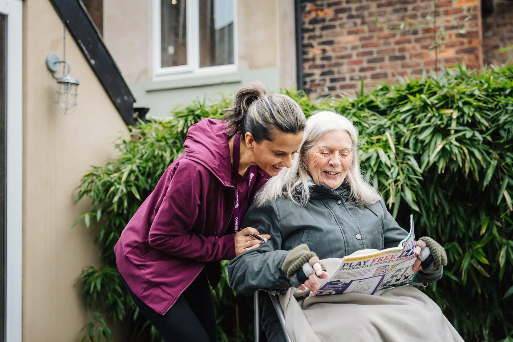 A caregiver smiles while assisting an older woman in a wheelchair who is reading a book outdoors. - Home Instead