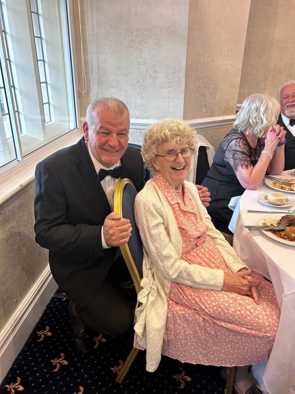 An elderly woman in a pink dress and white cardigan sits at a table, smiling with a man in a tuxedo kneeling beside her. - Home Instead