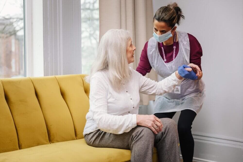 A caregiver assists an elderly woman sitting on a yellow sofa while holding her hand in a brightly lit room. - Home Instead