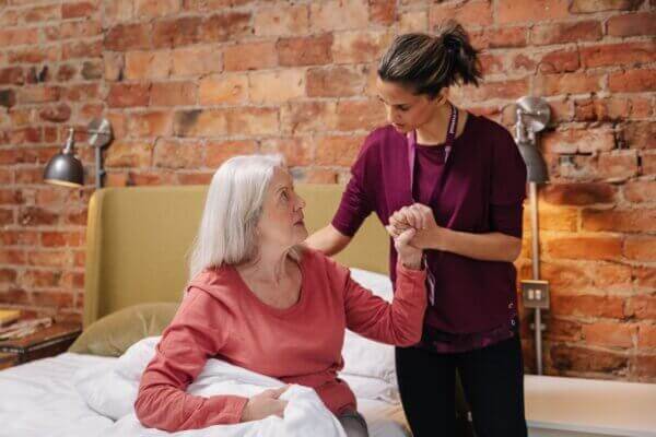 A caregiver assists an elderly woman with getting out of bed in a room with brick walls. - Home Instead