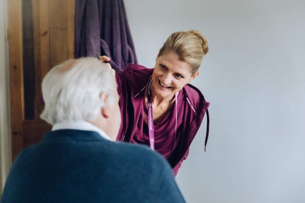 A caregiver with a name badge smiles and talks to an elderly person sitting, seen from behind, in a home setting. - Home Instead