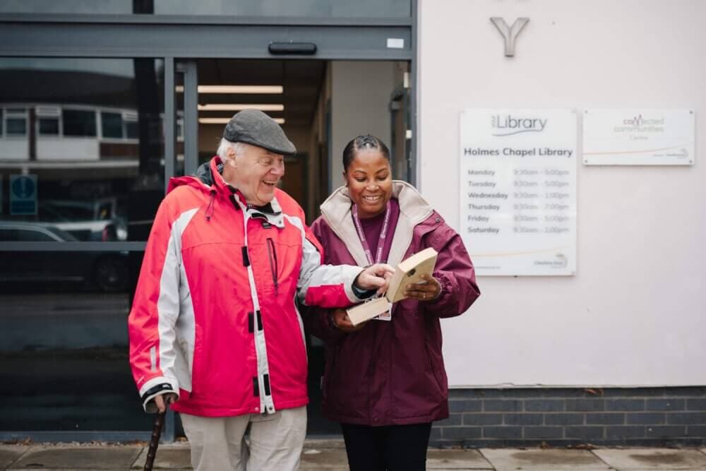 An older man and a younger woman smile while looking at a book outside Holmes Chapel Library. - Home Instead