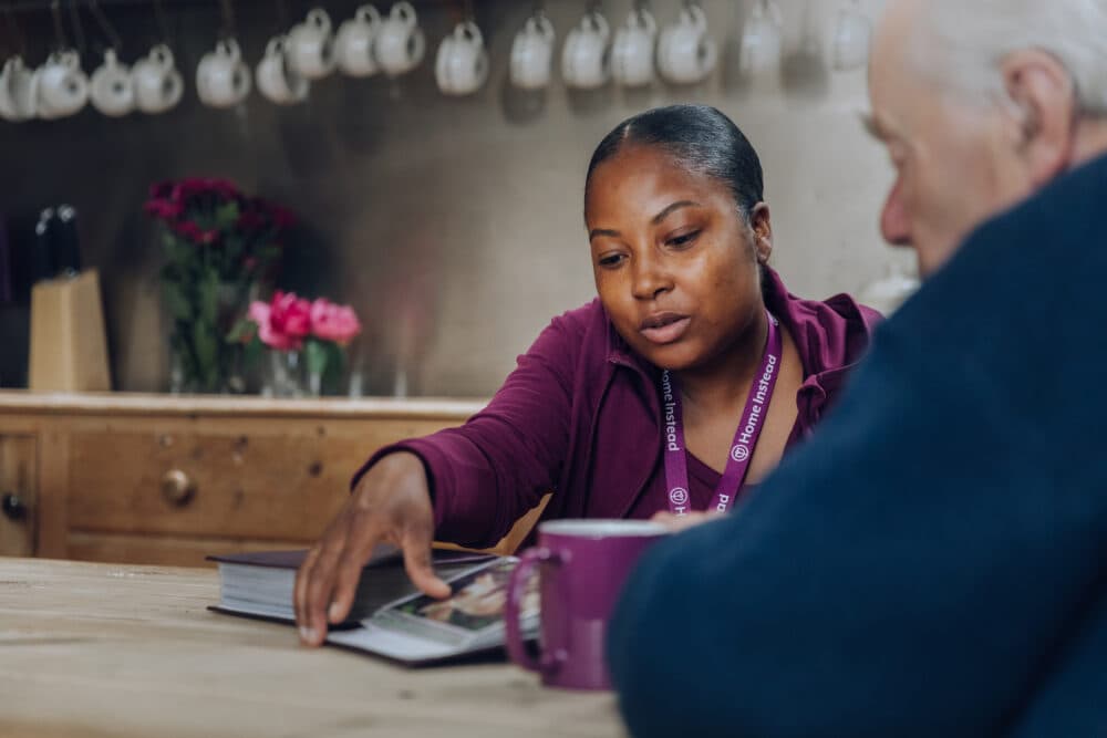 A woman wearing a lanyard showing a book to an older man in a kitchen with hanging cups and flowers in the background. - Home Instead