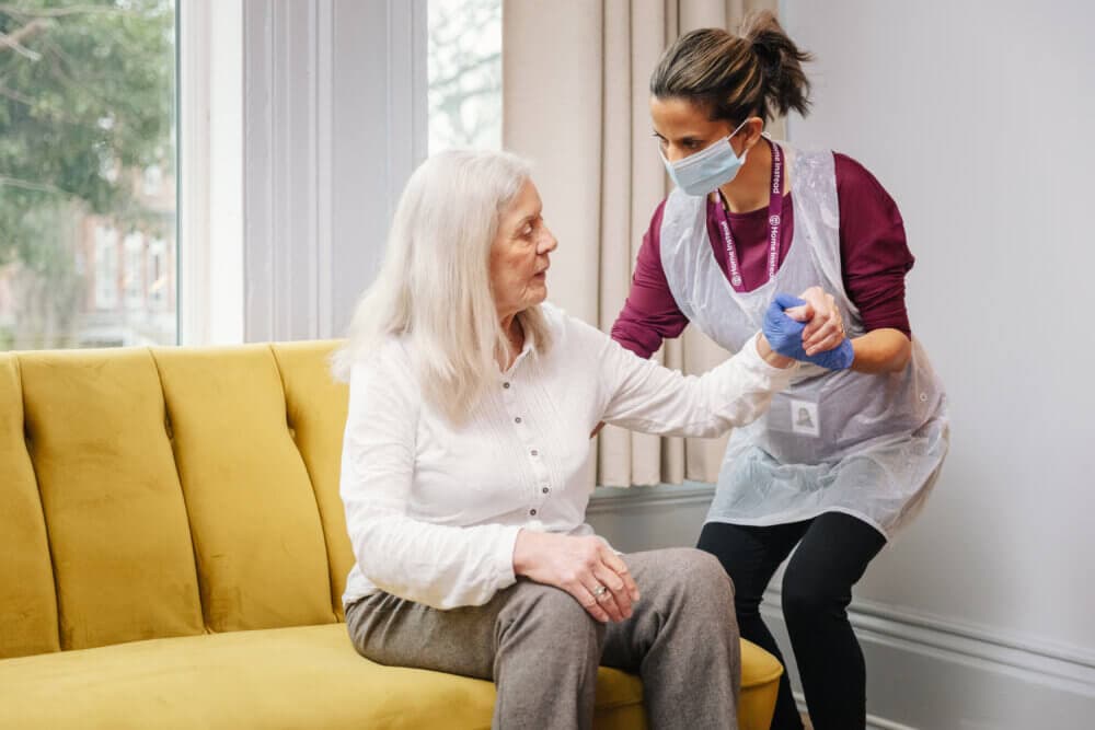 A caregiver assists an elderly woman to stand up from a yellow sofa in a well-lit room. - Home Instead