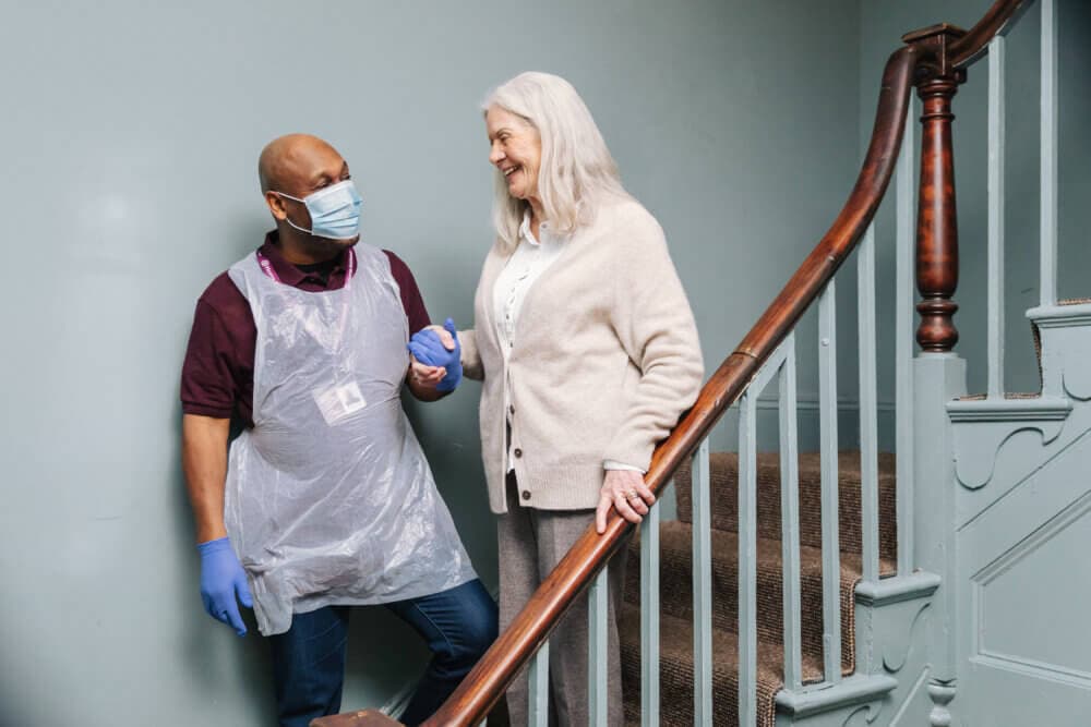 A caregiver in a mask and gloves helps an elderly woman walk down a staircase while holding her hand. - Home Instead
