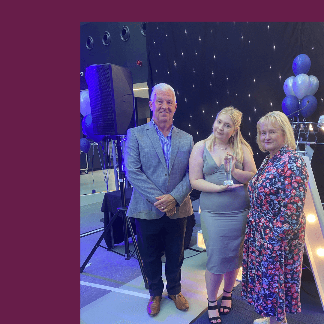 Three people stand together at an event, with a woman in the center holding a trophy and smiles all around. - Home Instead