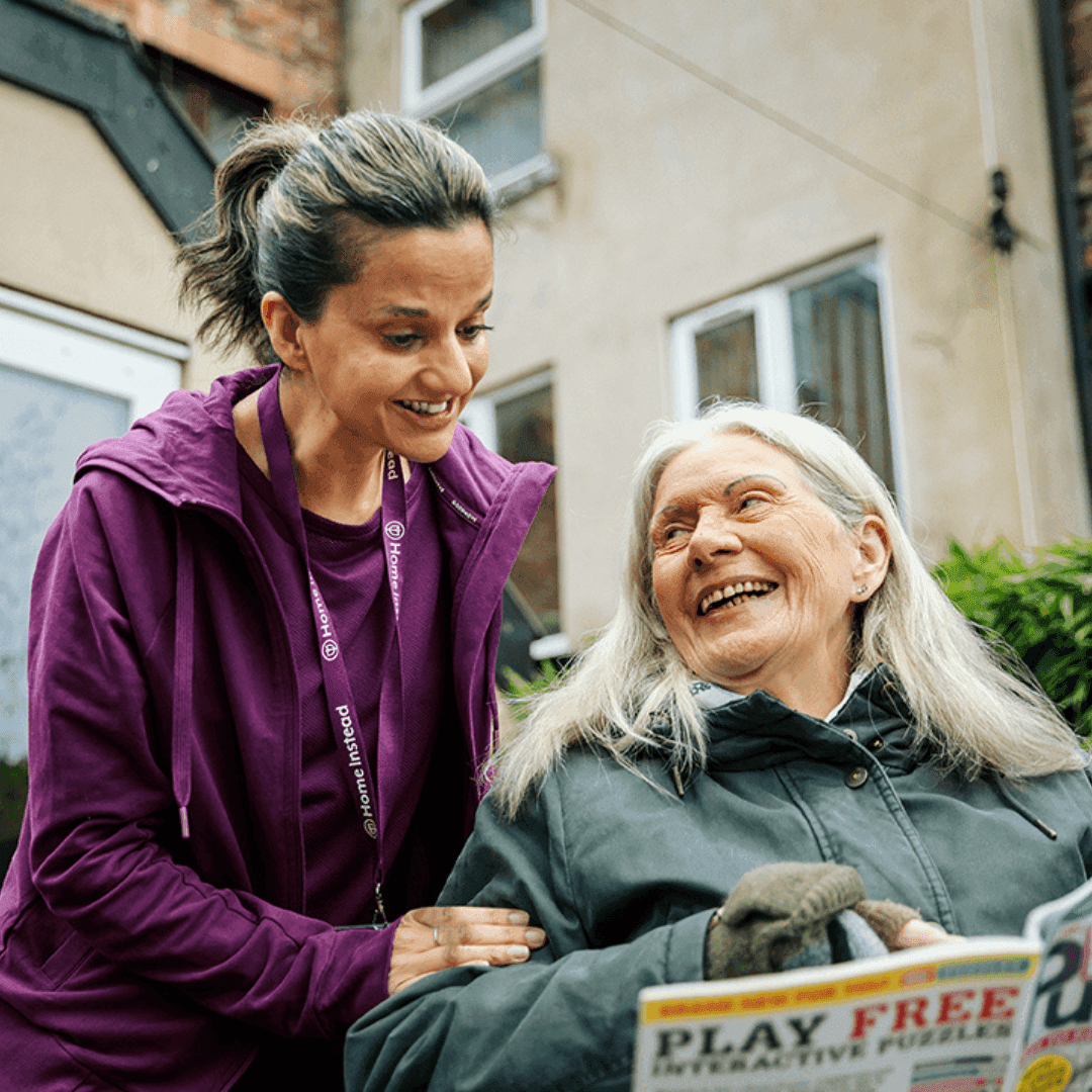 A caregiver in a purple jacket smiles at an elderly woman in a wheelchair who is also smiling. - Home Instead