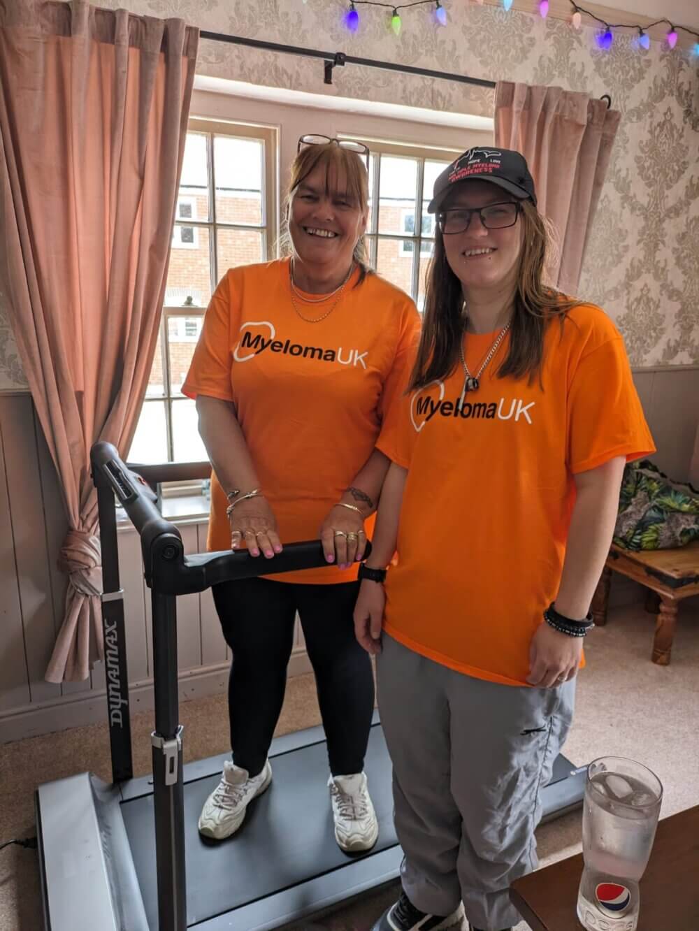 Two people wearing orange "Myeloma UK" shirts stand beside a treadmill indoors, smiling at the camera. - Home Instead