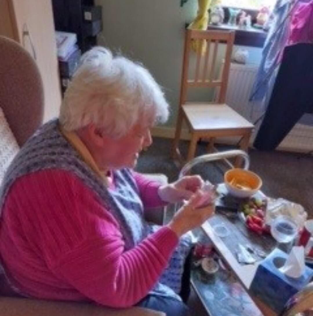 Elderly woman sitting in an armchair, knitting or handling yarn with various items on a table in front of her. - Home Instead