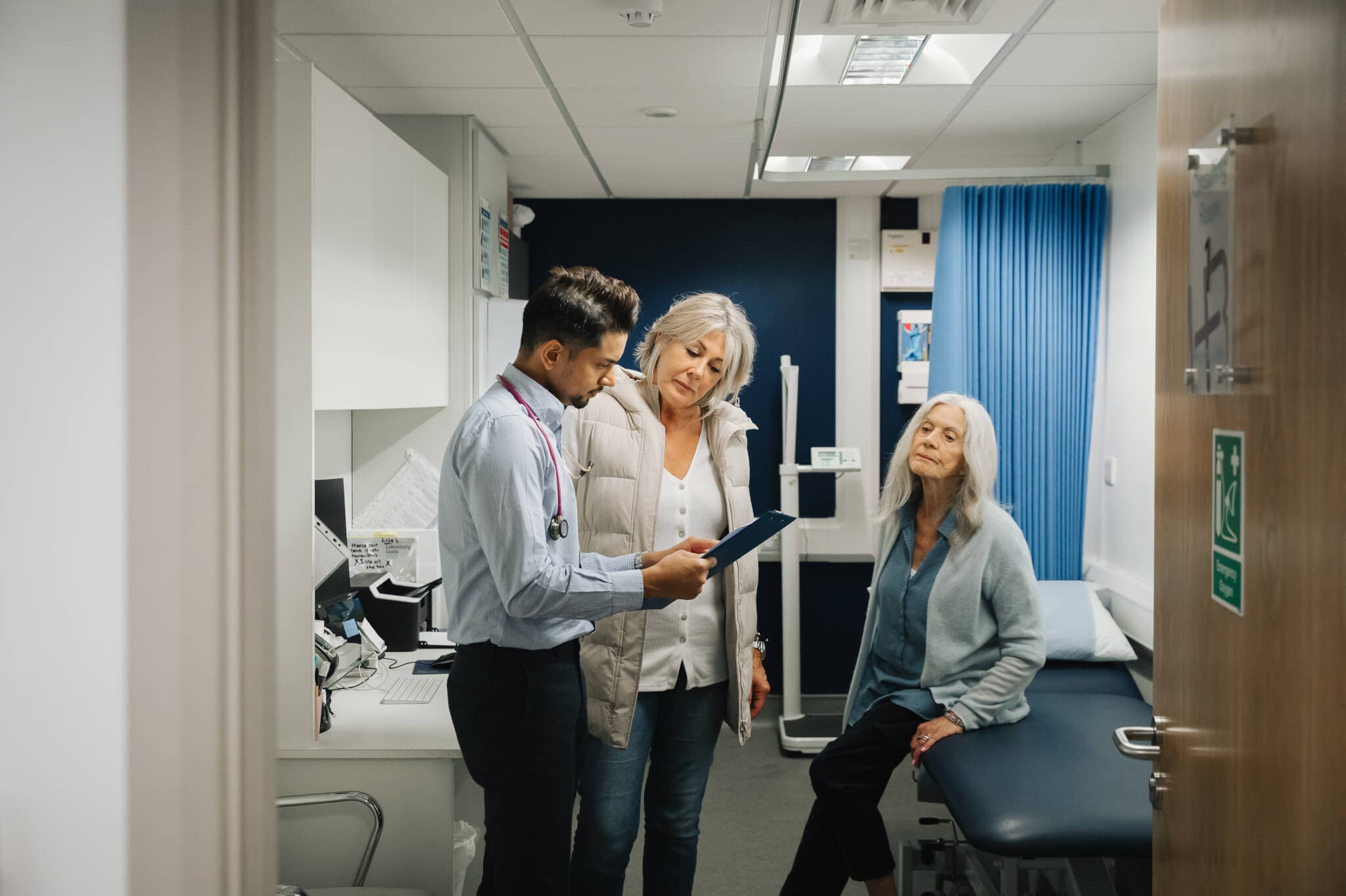 Doctor holding a clipboard and discussing with two women, one standing and one sitting on an examination table in a clinic. - Home Instead