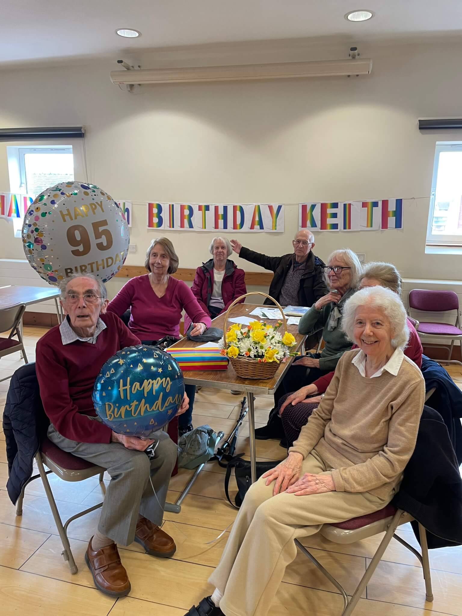 A group of elderly people at a 95th birthday celebration, with balloons and a "Happy Birthday" banner on the wall. - Home Instead