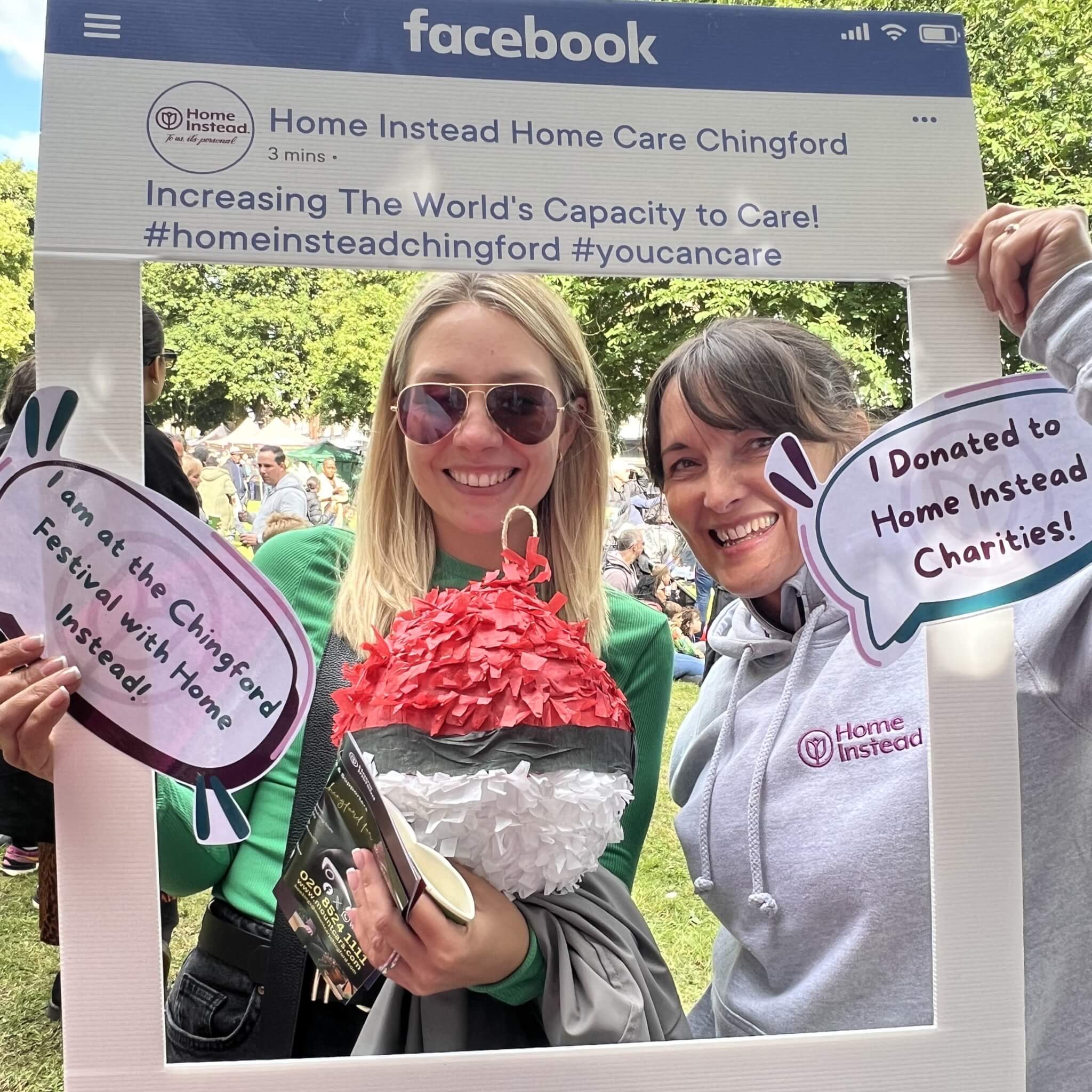 Two women smiling, holding signs related to Home Instead Charities at the Chingford Festival, posing with a Facebook frame prop. - Home Instead