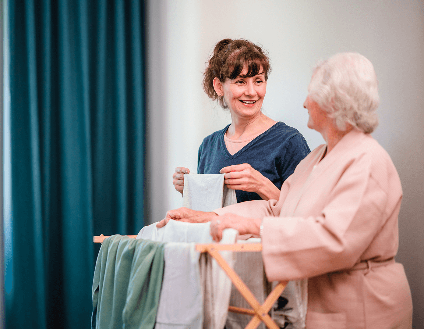 A young woman and an elderly woman smiling and folding laundry together in a cozy room. - Home Instead