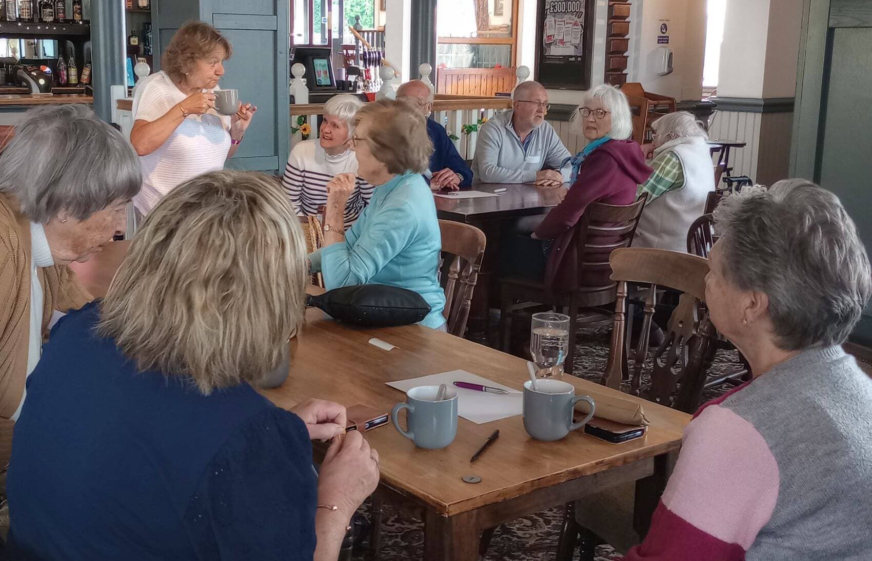A group of older adults sitting and standing around a coffee shop table, engaged in conversation. - Home Instead