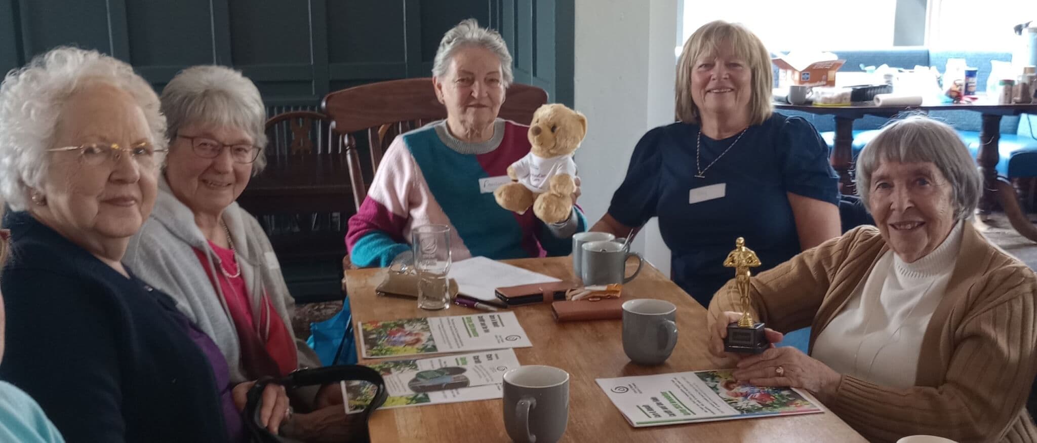 A group of older women smiling at a table with a teddy bear, mugs, and booklets in a cozy setting. - Home Instead