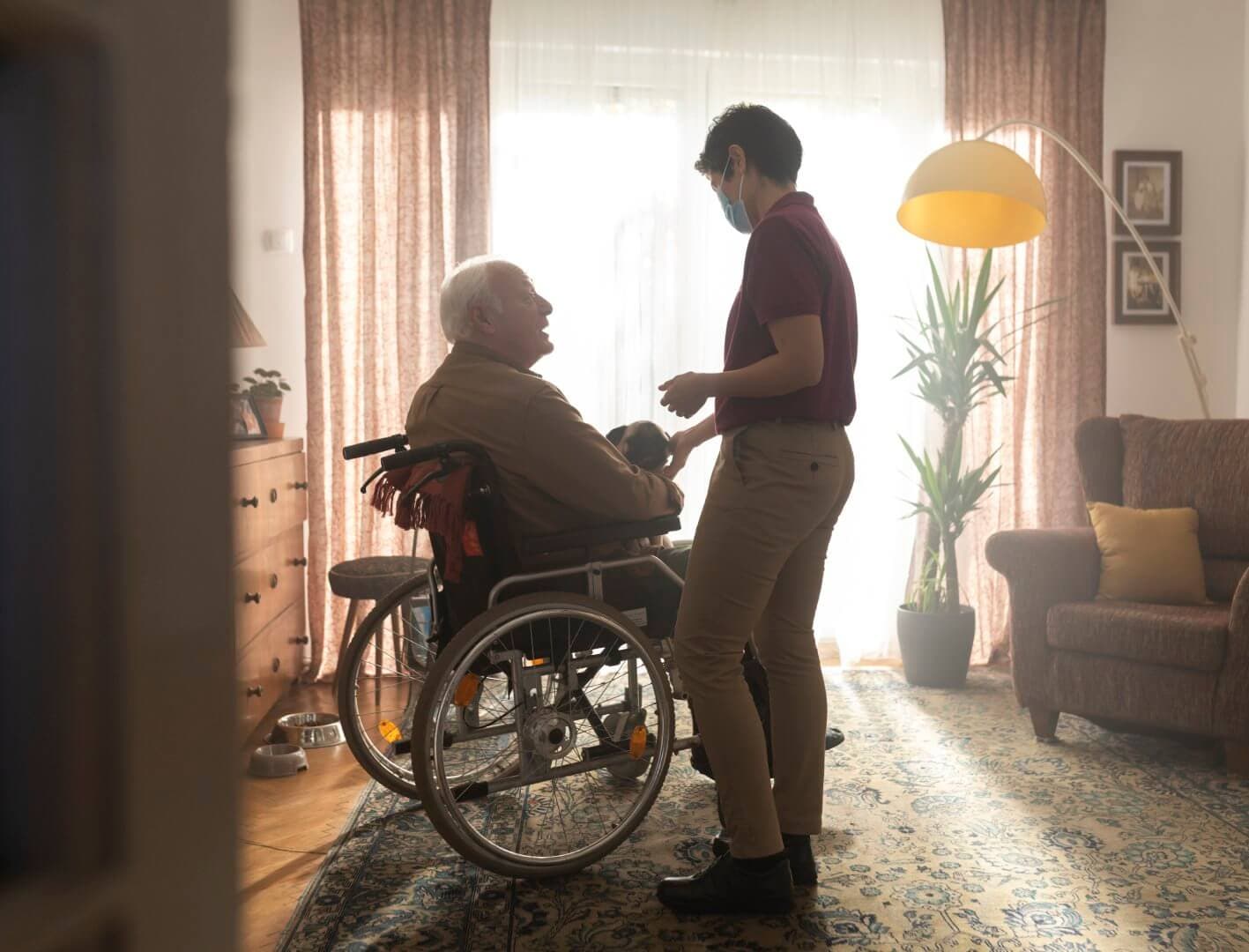 A caregiver wearing a mask assists an elderly man in a wheelchair in a cozy, sunlit living room. - Home Instead