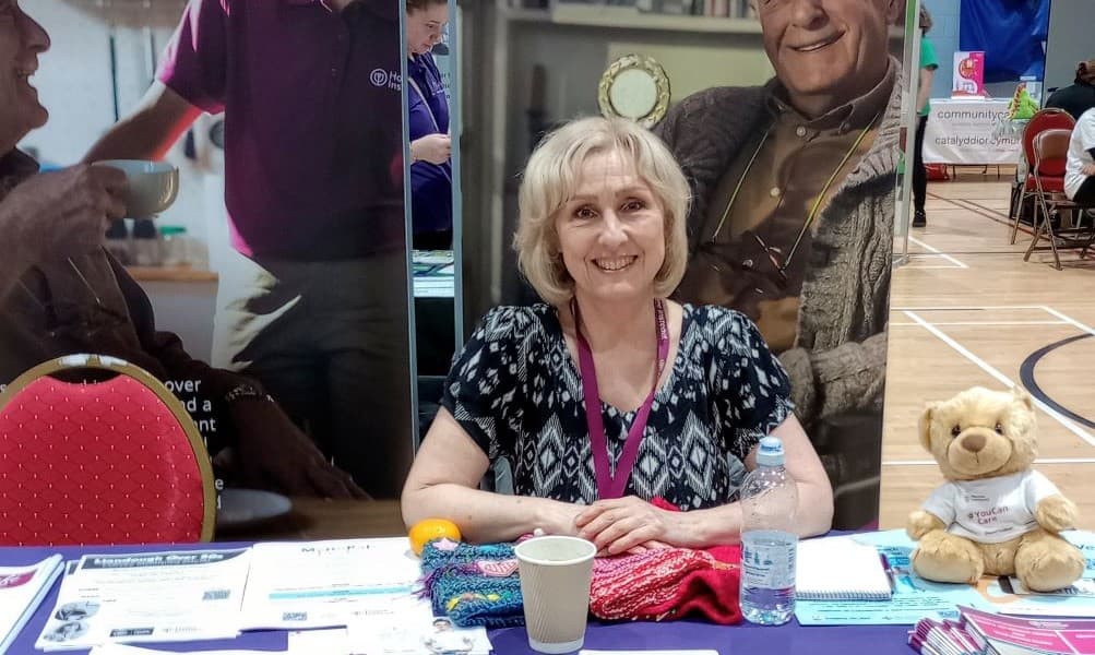 A woman with blond hair sits at a booth with brochures, a stuffed bear, and a water bottle in a gymnasium. - Home Instead
