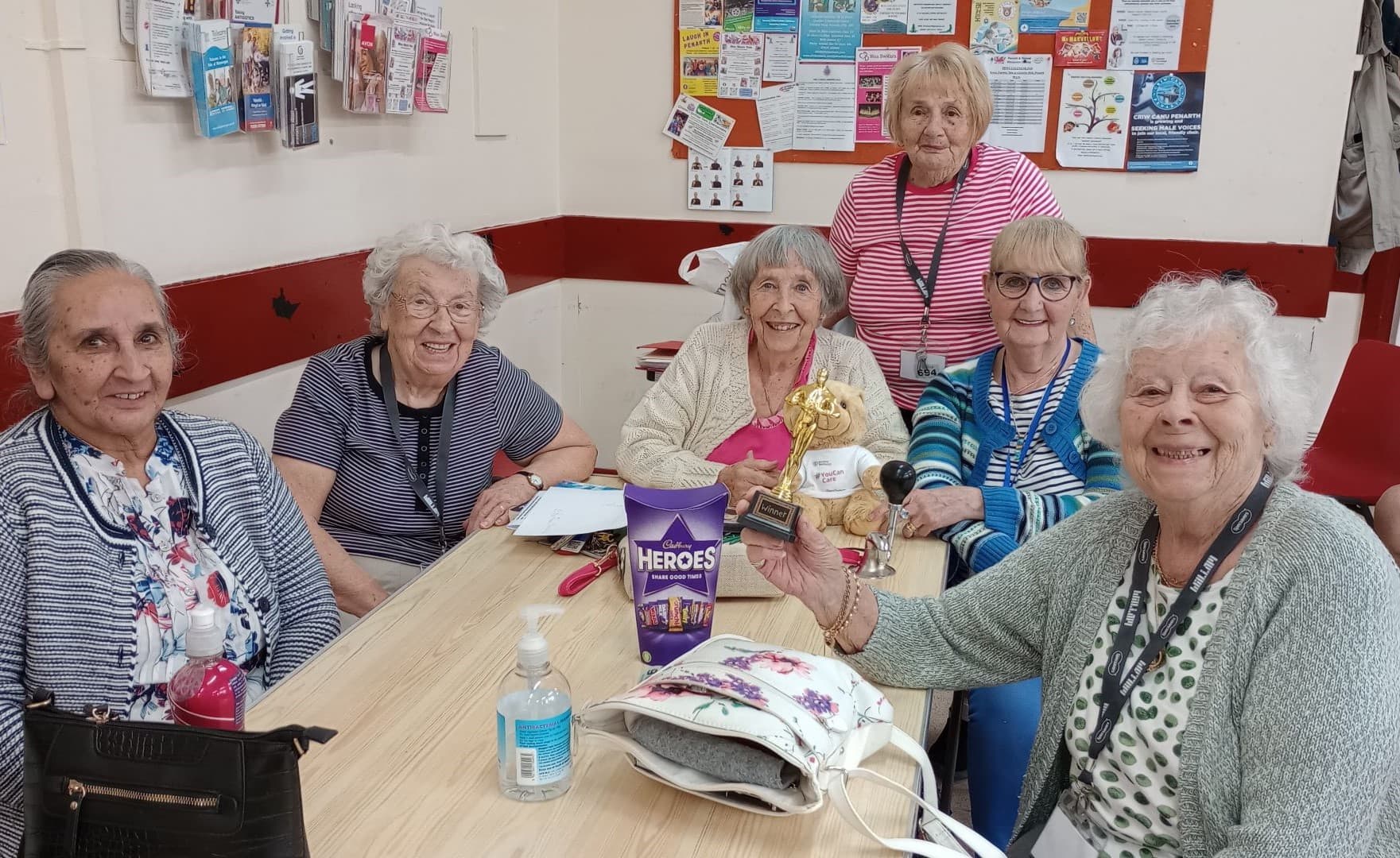 A group of elderly women seated around a table, smiling, with one holding a small trophy and another holding a stuffed lion. - Home Instead