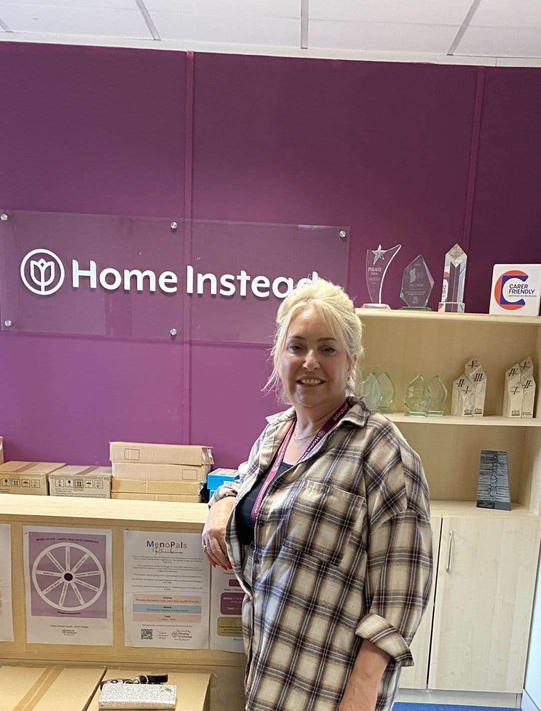 A woman standing by a reception desk with "Home Instead" sign and awards on a shelf in the background. - Home Instead