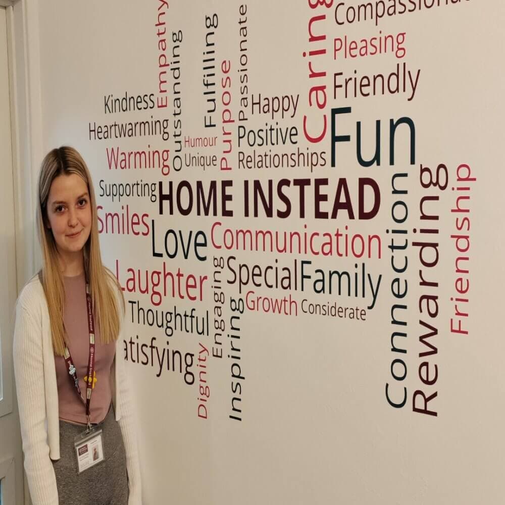 A woman stands in front of a wall covered with positive words such as "fun," "love," "family," and "rewarding. - Home Instead