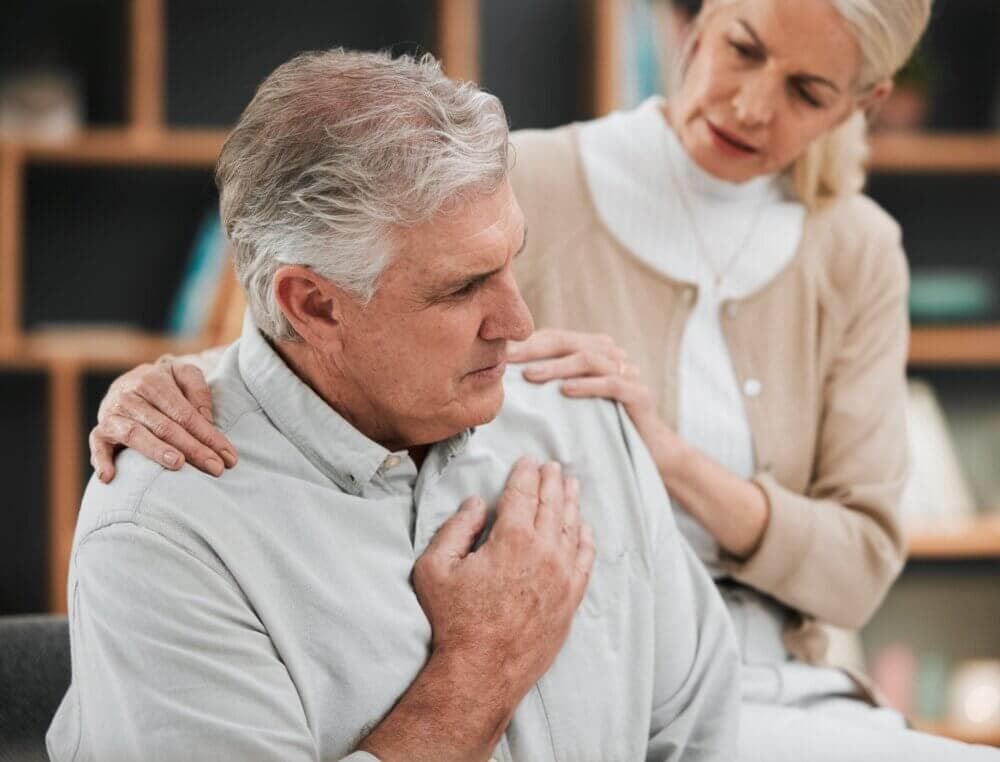 Elderly man clutching his chest in discomfort, while a concerned woman places her hands on his shoulders for support. - Home Instead