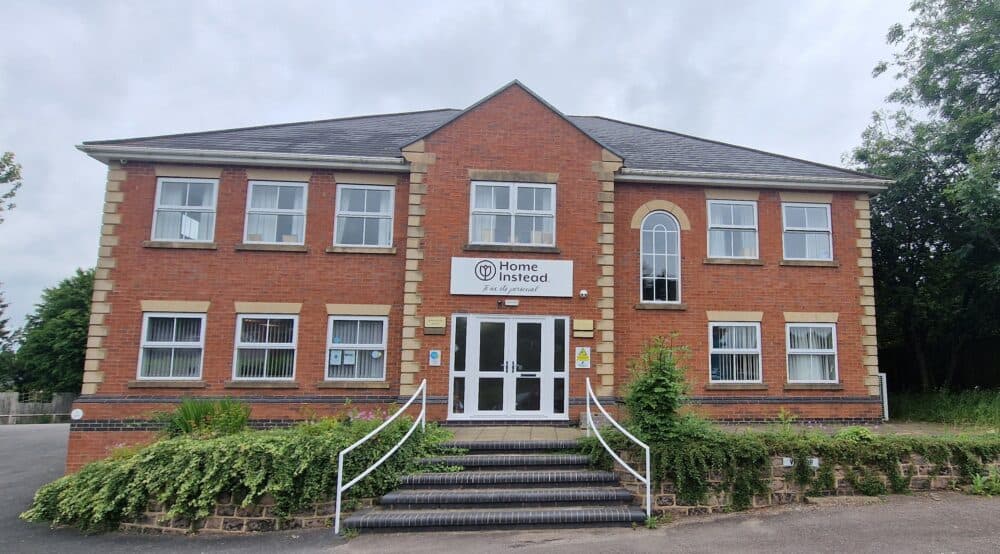 A two-story brick building with a sign reading "Home Instead" above the entrance, flanked by staircases and greenery. - Home Instead