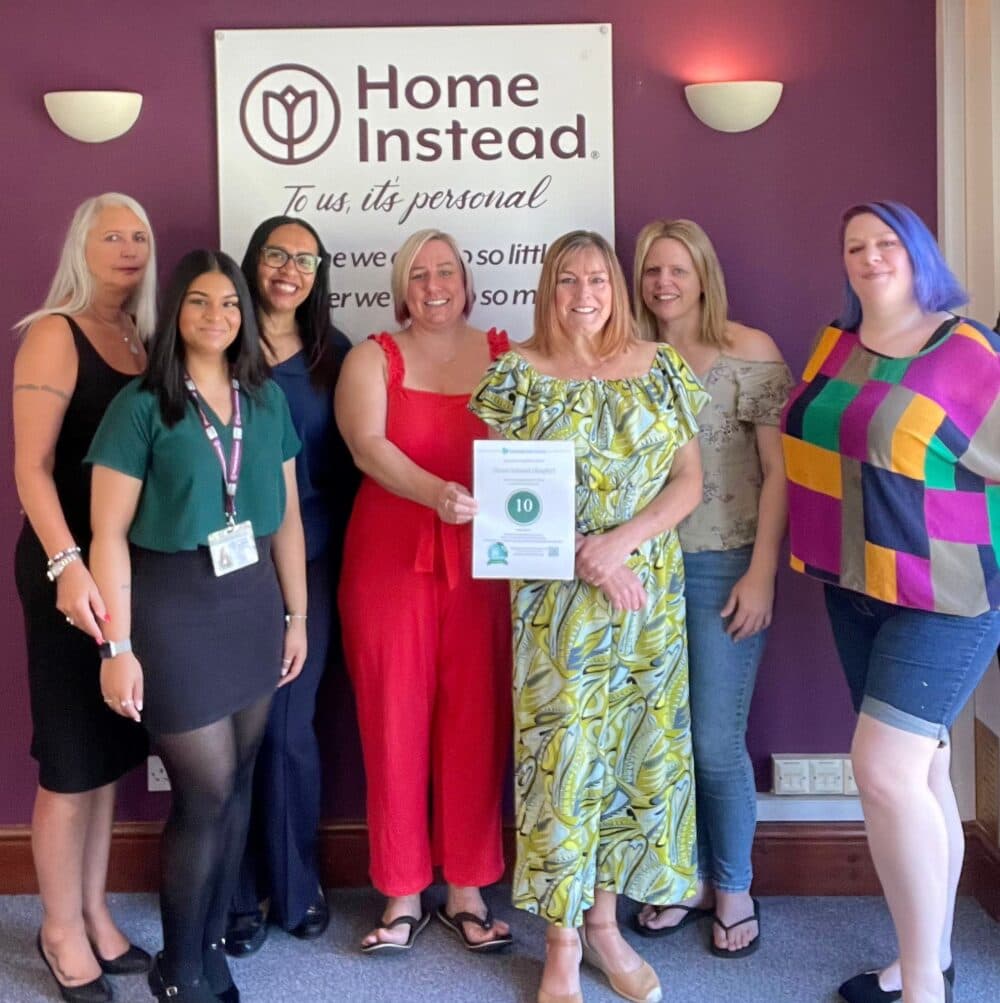 A group of seven women standing together in front of a "Home Instead" sign, with one holding an award certificate. - Home Instead