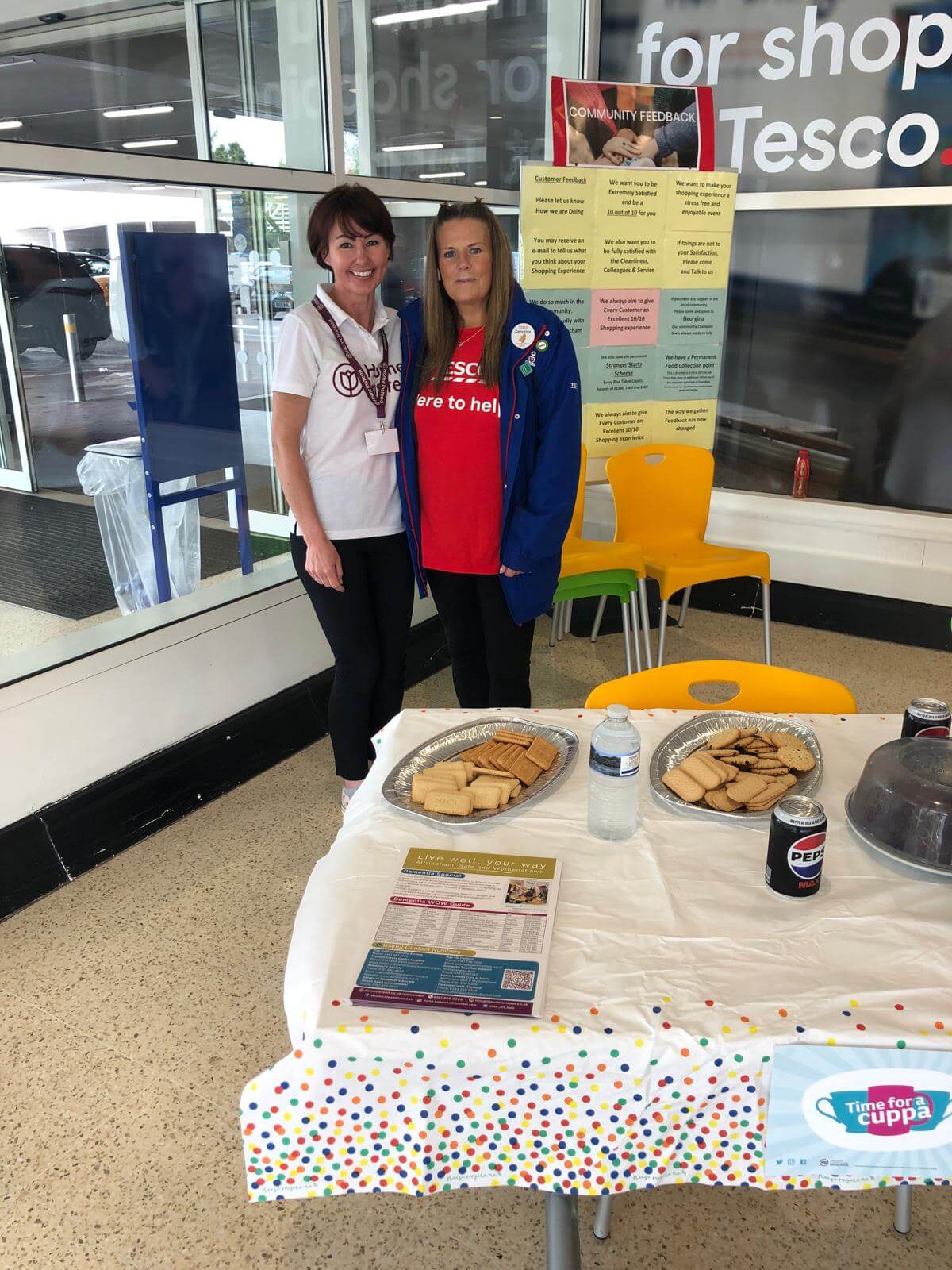 Two women stand behind a table with snacks and flyers at a Tesco store entrance, participating in a community event. - Home Instead