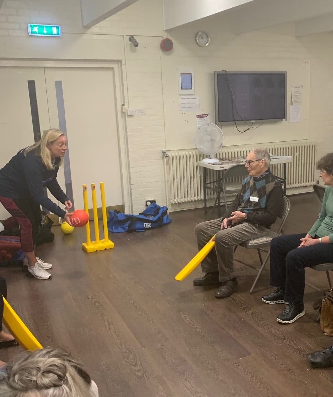 A woman helps an older man seated in a chair with a yellow bat participate in indoor cricket with others watching. - Home Instead