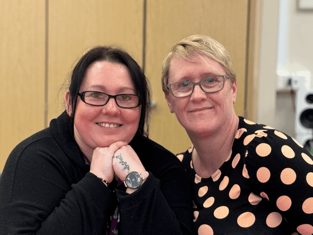 Two people smiling, posing closely together. One wears glasses and a watch, the other wears a polka dot shirt. - Home Instead