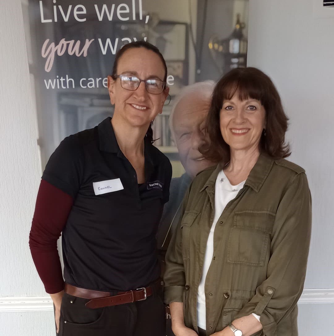 Two women standing and smiling in front of a banner with text. One is in a dark outfit, and the other is in a green jacket. - Home Instead