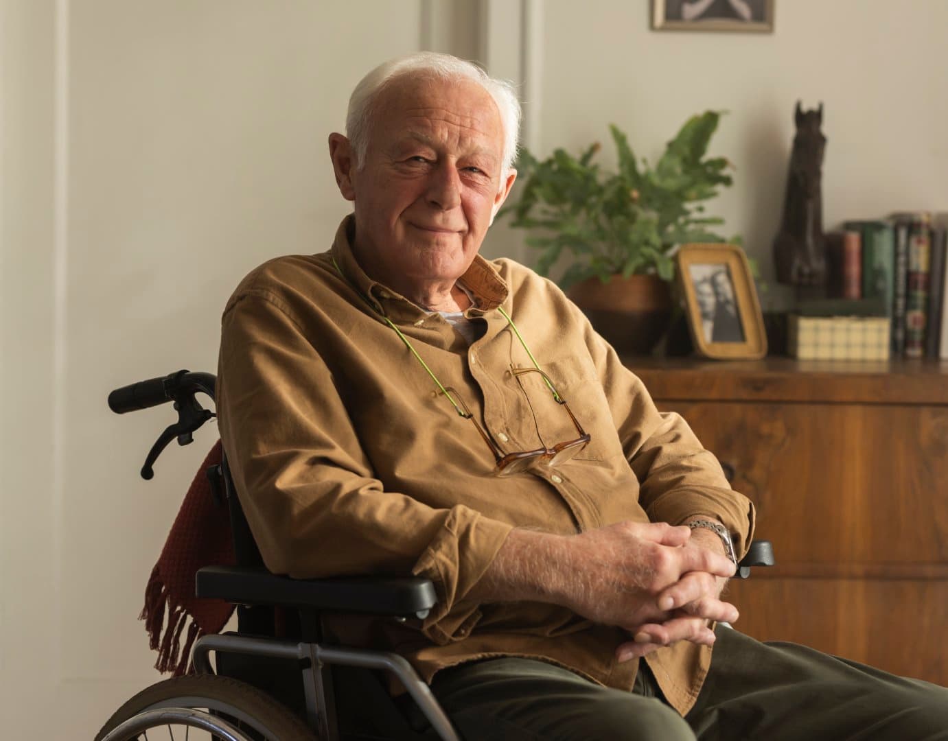 An elderly man with white hair sits in a wheelchair, smiling, with a bookshelf and plant in the background. - Home Instead