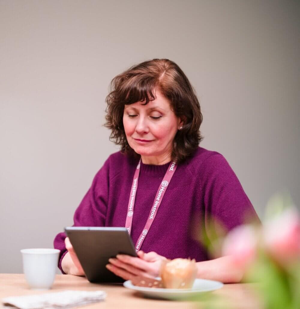 Woman in a purple top reads on a tablet at a table with a cup and a plate, blurred flowers in the foreground. - Home Instead