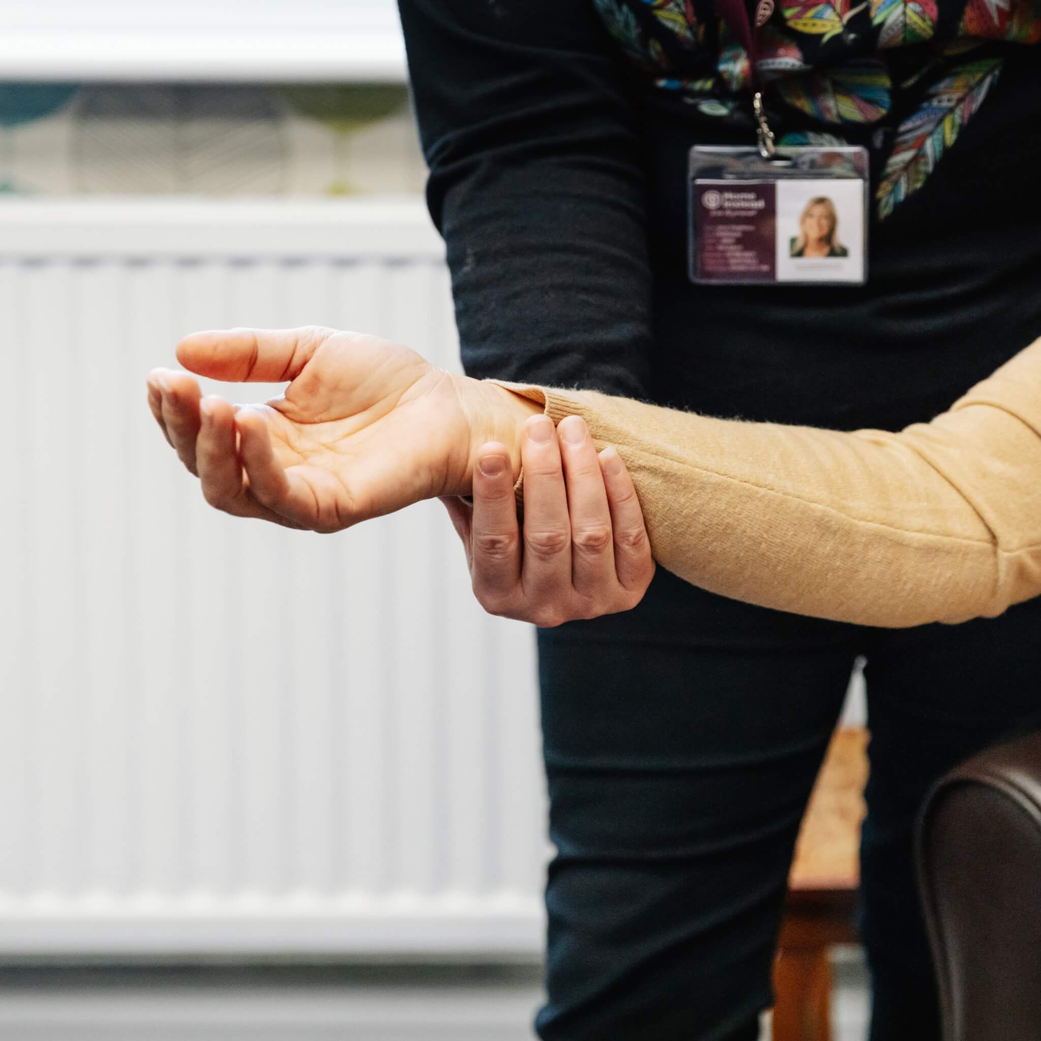 A Home Instead Care Professional in a black shirt guides the arm of another person wearing a beige sweater who is seated.