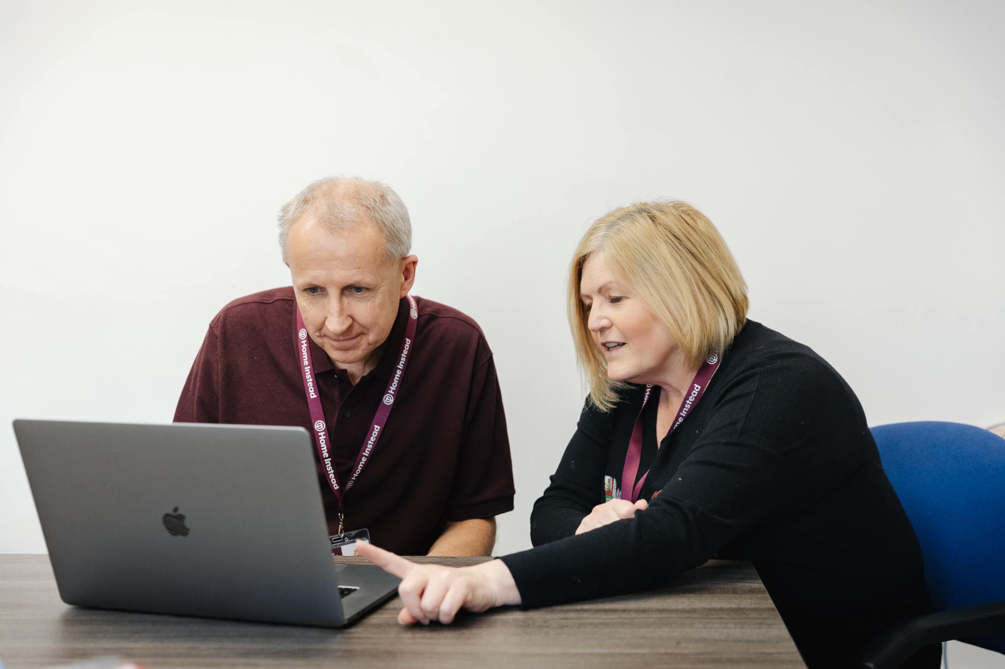 Two people wearing lanyards sit at a desk, collaborating and looking at a laptop screen. - Home Instead