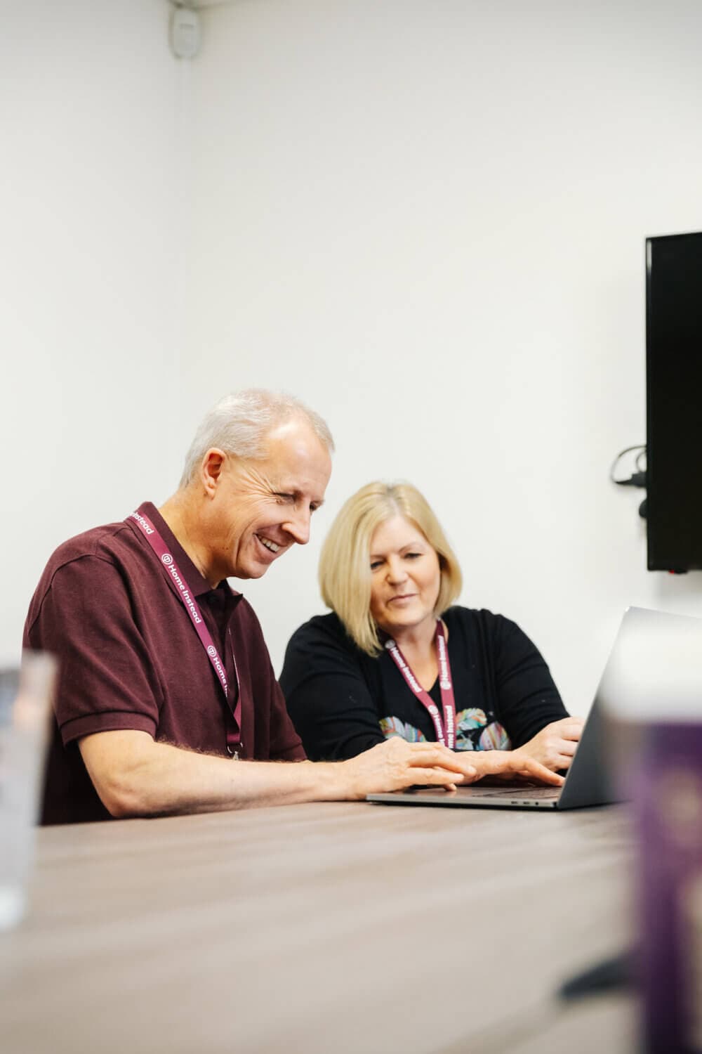 Two people sitting at a table, smiling and working on a laptop. They are in a bright office space. - Home Instead