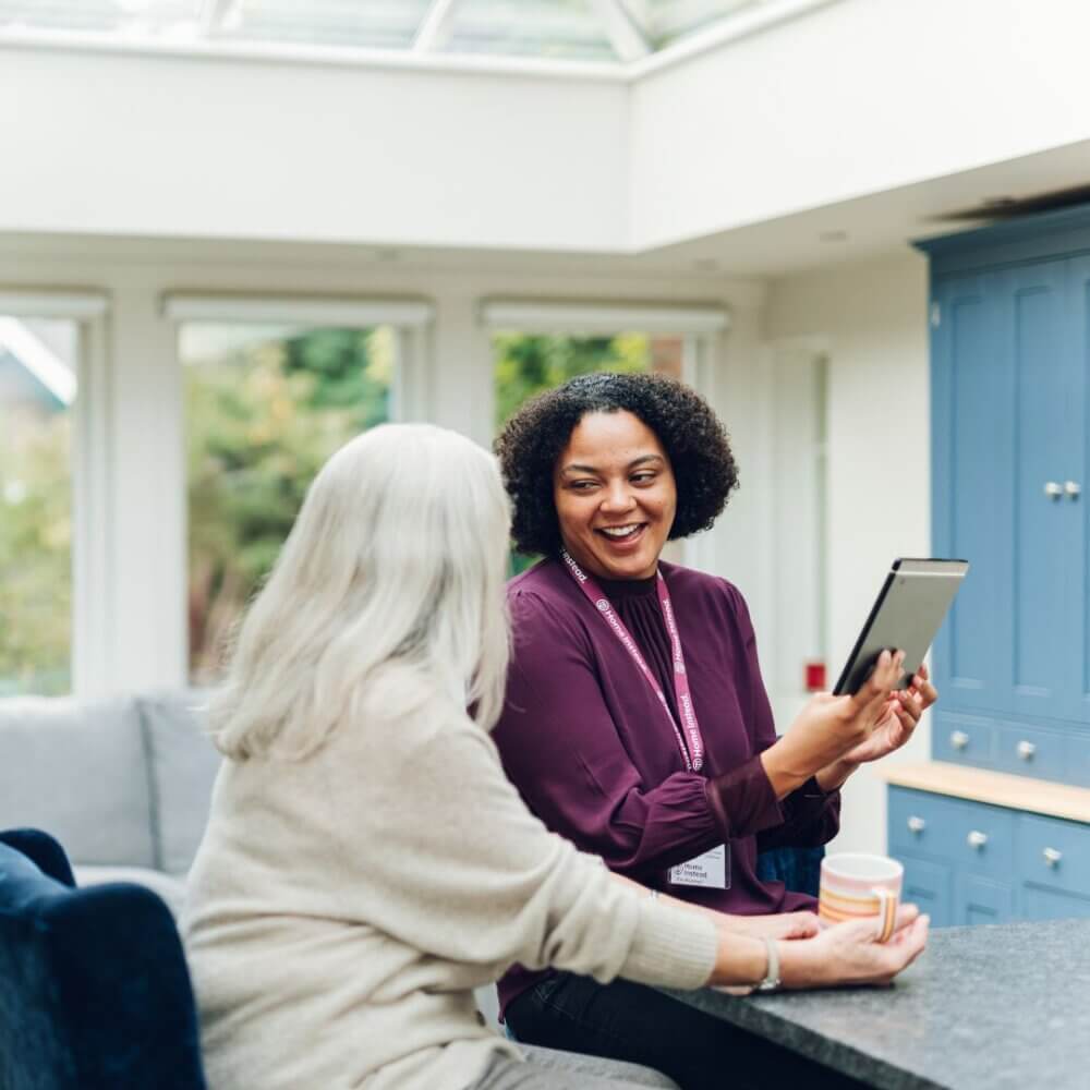 Two people sitting in a bright kitchen, smiling and looking at a tablet together. - Home Instead
