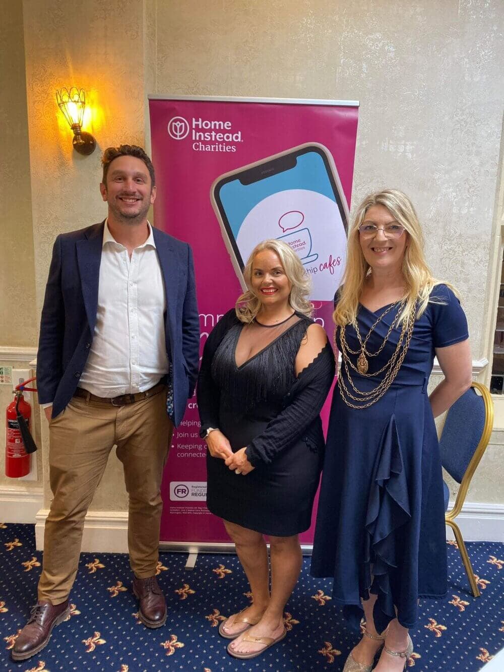 Three people standing in front of a "Home Instead Charities" banner in a room with a chandelier and patterned carpet. - Home Instead