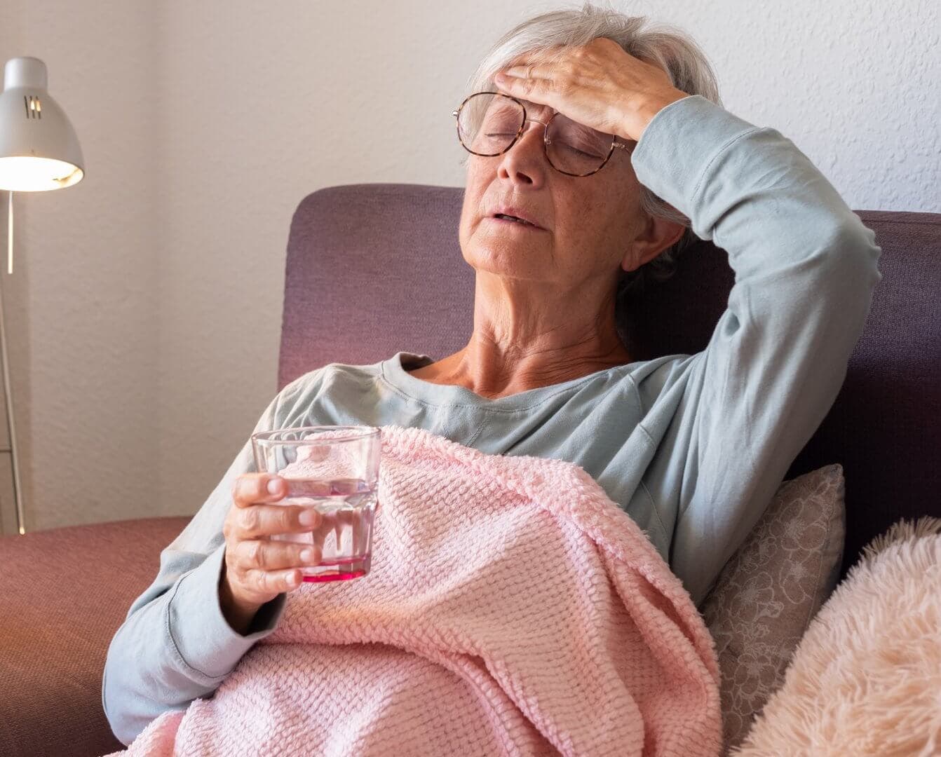 Elderly woman with glasses holding a glass of water, resting on a couch with a pink blanket, appearing unwell. - Home Instead