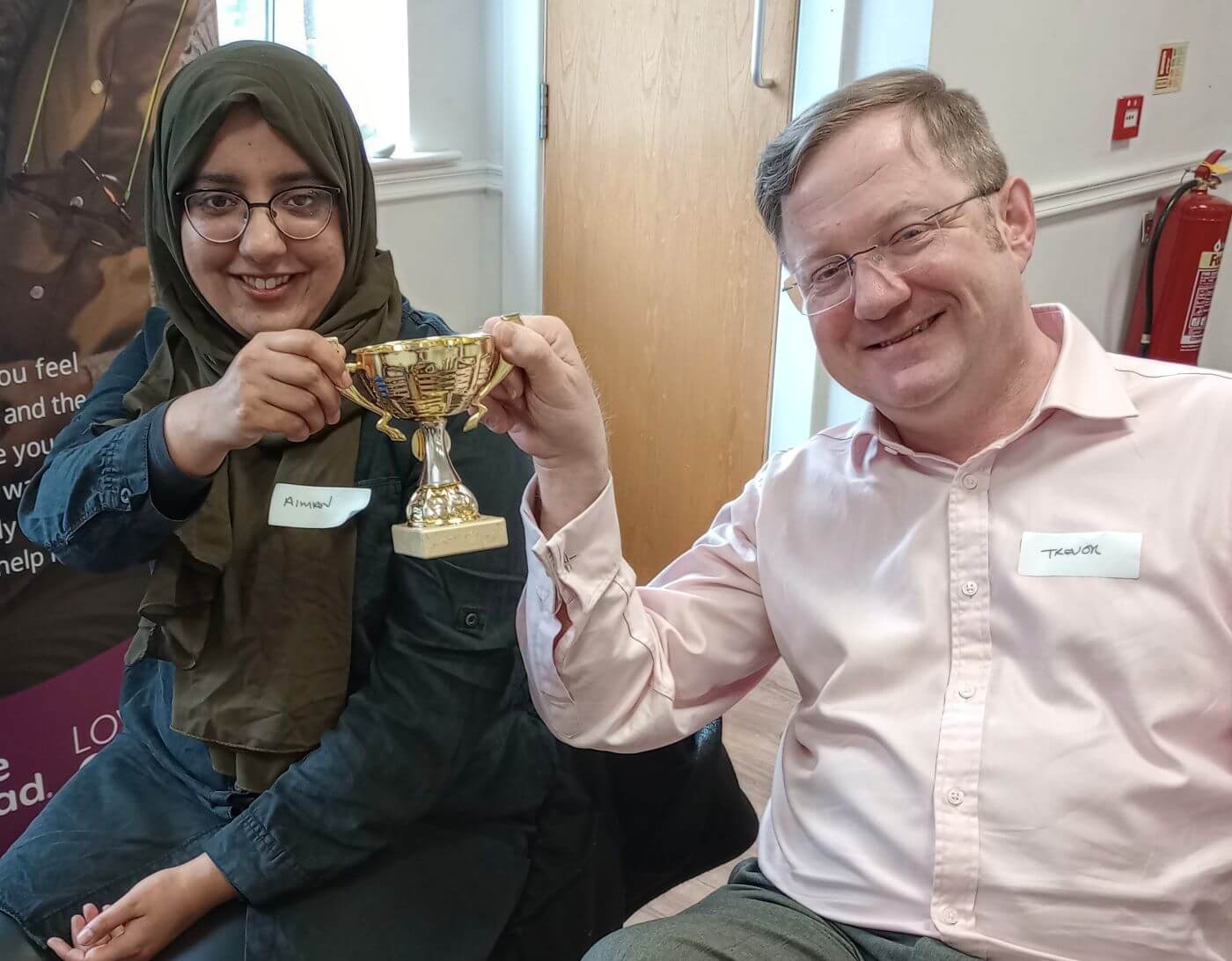 Two people smiling and holding a gold trophy together; they are seated indoors and wearing name tags. - Home Instead
