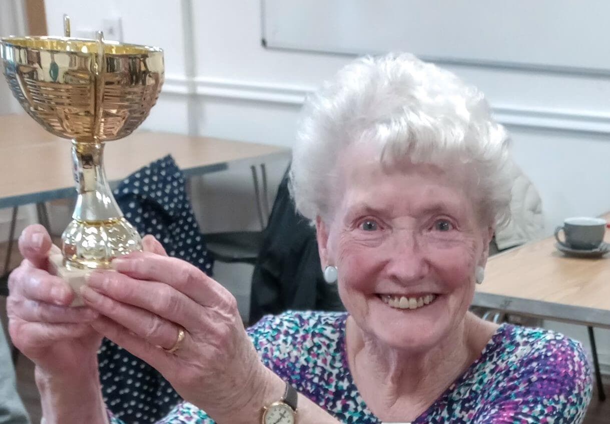 Smiling elderly woman holding a golden trophy in a classroom setting. - Home Instead