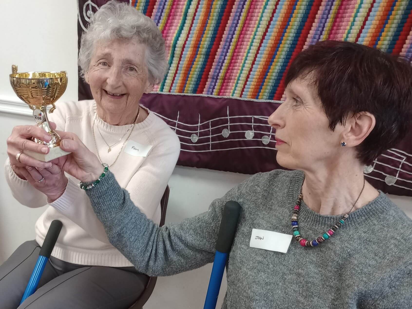 Two women smiling and holding up a gold trophy, seated with a colorful tapestry behind them. - Home Instead