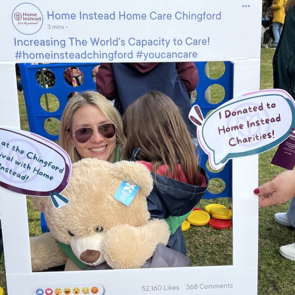 Person holding a large teddy bear and hugging a child, posing with a photo frame at an outdoor event. - Home Instead