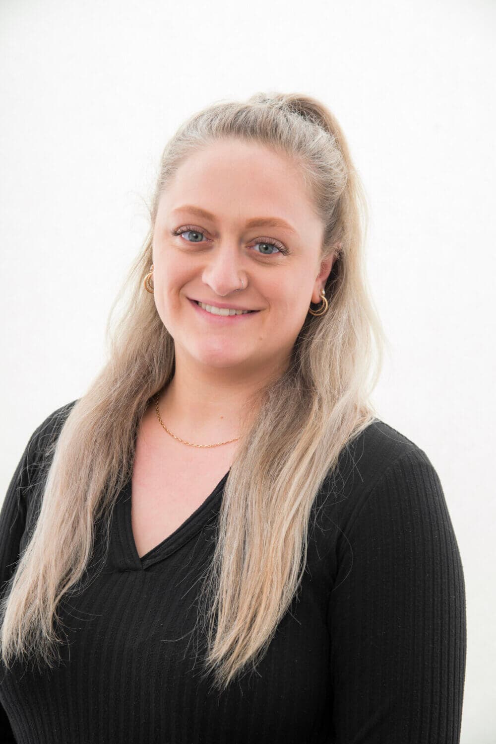 A woman with long blonde hair in a ponytail, wearing a black top, smiles at the camera. - Home Instead