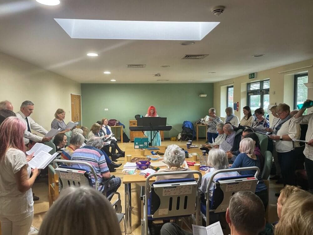 A group of people, some seated and others standing, singing together in a well-lit room. A conductor with red hair leads them. - Home Instead