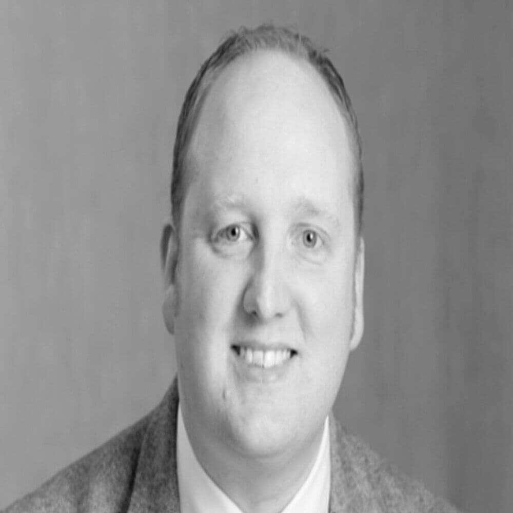 Black and white portrait of a smiling man in a suit jacket and tie against a plain background. - Home Instead