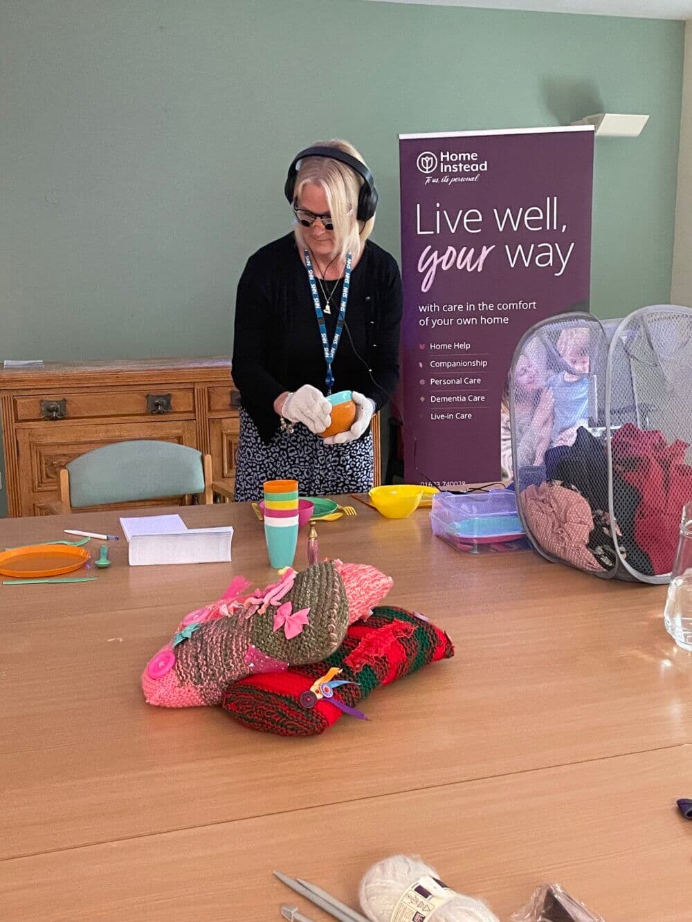 A woman with gloves and headphones prepares a meal in a kitchen with a banner reading, "Live well, your way. - Home Instead
